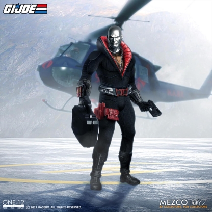 "Mezco One:12 Collective Destro action figure standing in front of a blurred helicopter, holding his Cobra-issued briefcase in one hand and a pistol in the other, on what appears to be a helipad."