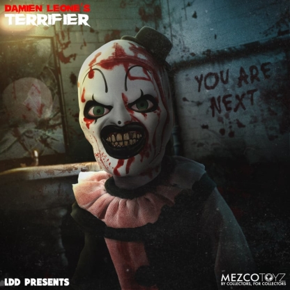 A close-up of the Living Dead Doll of Art the Clown, showing his blood-splattered white face, top hat, green eyes, and exposed rotten teeth. In the blurry background, a dark, blood-stained wall has "YOU ARE NEXT" written on it.