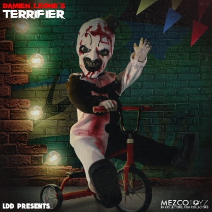 The Living Dead Doll of Art the Clown is sitting on a red tricycle, with one leg lifted. He wears his blood-splattered clown suit and top hat, with a menacing grin and green eyes. The background is a brick wall with string lights and colorful triangular flags.